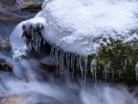 Eiszapfen und dicke Eisfigur an moosbedecktem Stein am Bachufer